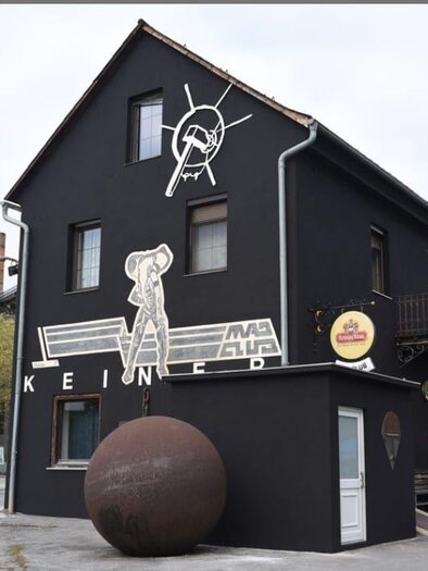 Ein schwarzes Gebäude mit einer auffälligen Wandmalerei. Vor dem Haus steht eine große, runde Skulptur und es sind einige Menschen zu sehen. | © keine Kenntnis