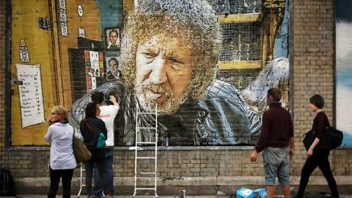 Eine große Wandmalerei eines Mannes mit lockigem Haar. Mehrere Menschen stehen davor und beobachten, während jemand daran arbeitet. | © keine Kenntnis