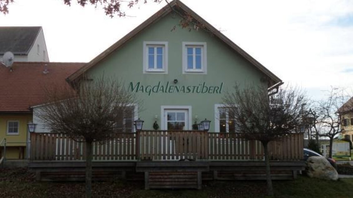 Ein charmantes Haus mit einem Holzsteg und zwei kleinen Bäumen vorne. Über dem Eingang steht der Schriftzug "Magdalena's Stüberl".