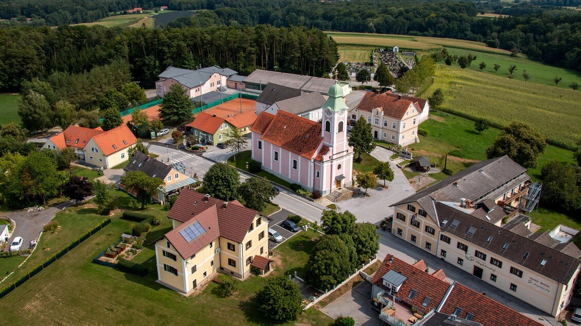 Eine ruhige Dorfansicht mit bunten Häusern und einer Kirche im Mittelpunkt. Umgeben von Feldern und Wäldern bietet die Landschaft eine idyllische Atmosphäre.