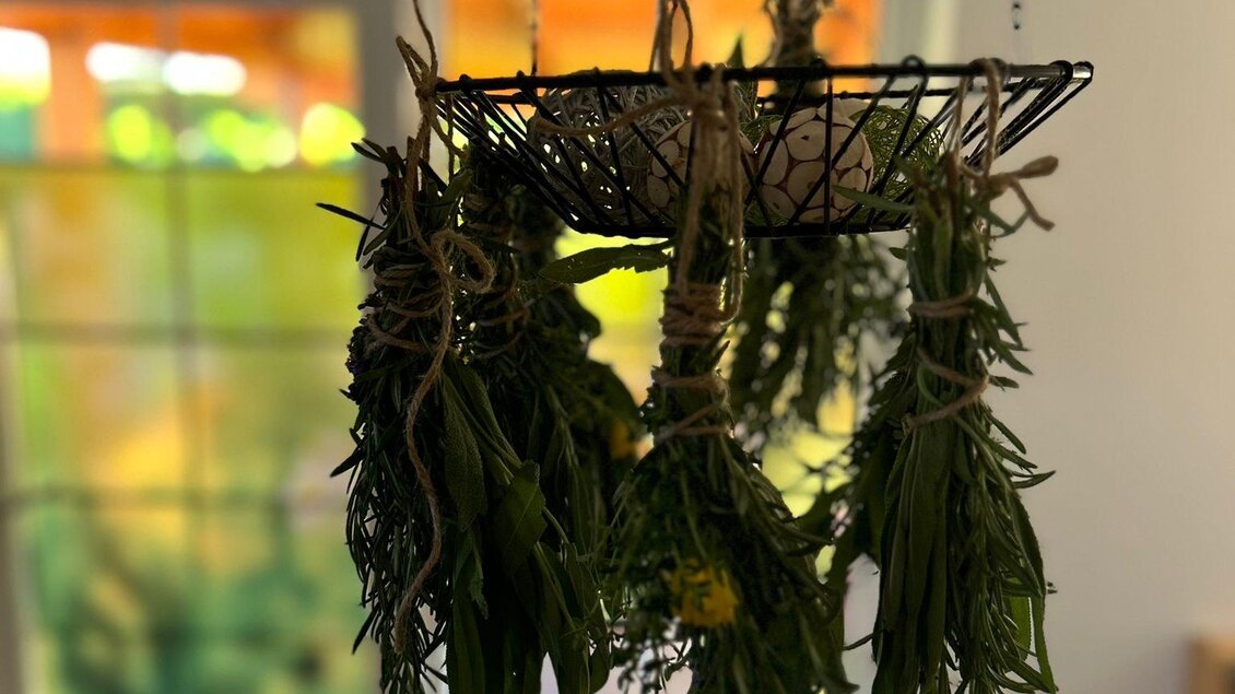 Ein Drahtkorb hängt von der Decke und enthält frisch gefüllte Kräuter. Im Hintergrund sind sanfte Farben und ein Fenster sichtbar.