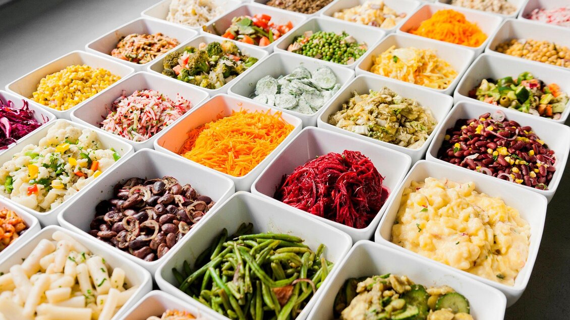 Eine Vielzahl von bunten Salaten und Beilagen in weißen Schalen. Die Anordnung zeigt eine große Auswahl an frischen Zutaten. | © Mangolds