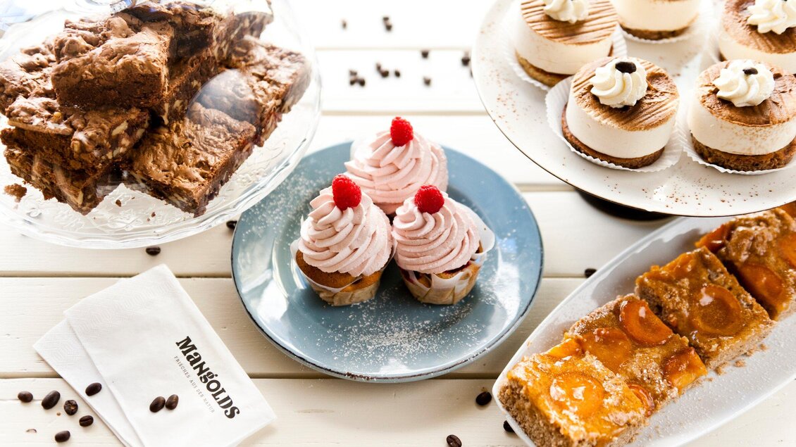 Eine Auswahl an köstlichen Desserts, darunter Cupcakes mit rosa Frosting, Torten und Schokoladenbrownies. Die süßen Leckereien sind ansprechend auf einem Tisch angerichtet. | © Mangolds