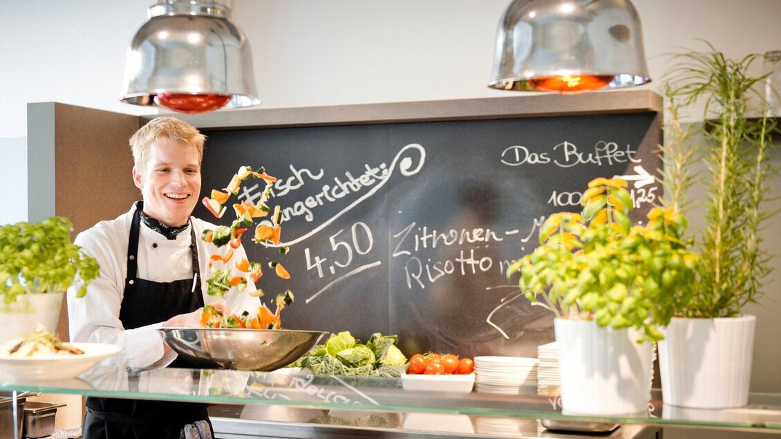 Ein Koch steht an einer modernen Küche und lächelt. Im Hintergrund sind frische Zutaten und eine Tafel mit Angeboten zu sehen. | © Mangolds