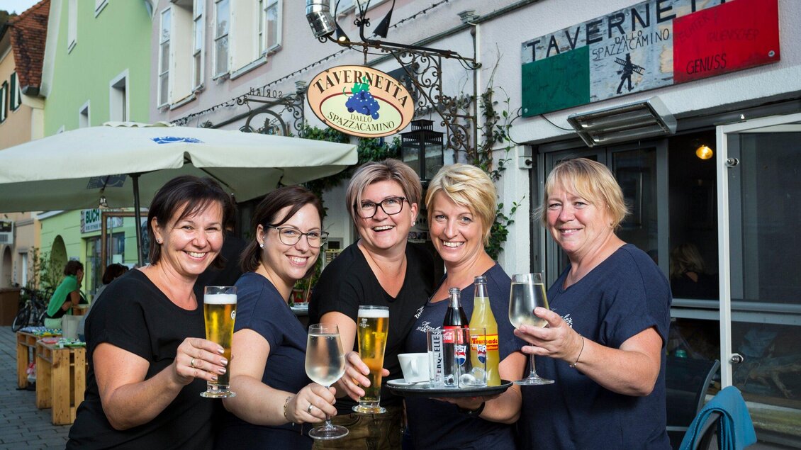 Fünf freundliche Frauen stehen vor einem Restaurant und halten Getränke in der Hand. Im Hintergrund ist das Schild des Restaurants "Taverne" zu sehen. | © TV Region Graz - René Vidalli