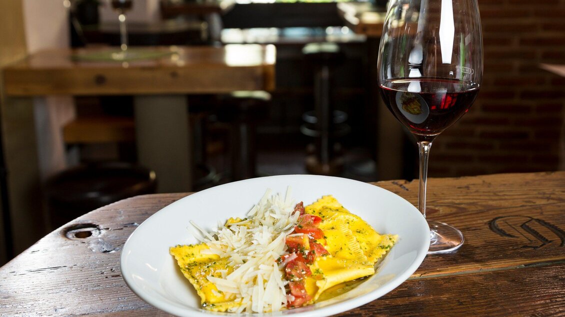 Ein Teller mit frischen Ravioli, garniert mit Tomaten und Parmesan. Daneben steht ein Glas Rotwein auf einem Holztisch. | © TV Region Graz - René Vidalli
