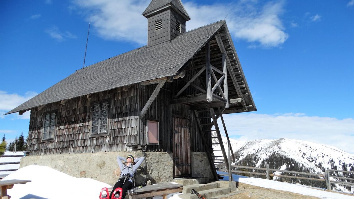 Eine traditionelle Holzhütte in schneebedeckter Landschaft. Im Vordergrund ist eine Person, die auf einem Schlitten sitzt und die Sonne genießt. | © Weges