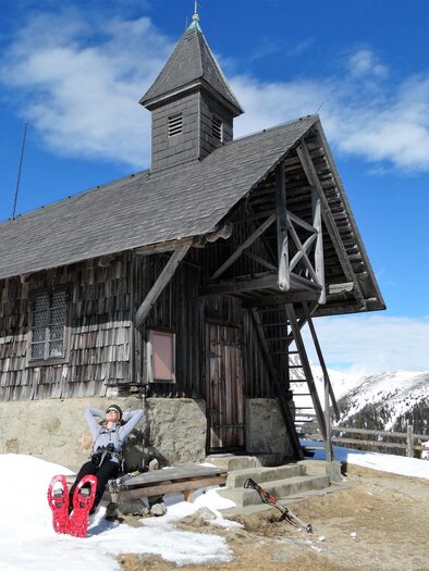 A traditional wooden cabin in a snow-covered landscape. In the foreground, there is a person sitting on a sled, enjoying the sun. | © Weges