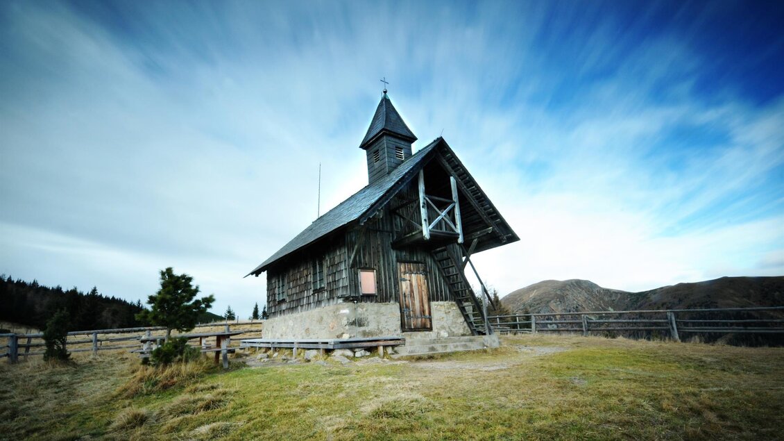 Eine alte Holzkirche auf einer Wiese mit sanften Hügeln im Hintergrund. Der Himmel ist blau mit bewegten Wolken. | © Eberhard