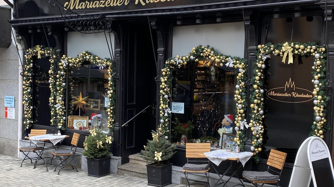 Ein gemütlicher Käseladen mit festlicher Dekoration. Tische und Stühle sind vor dem Laden aufgebaut, umgeben von grünem Rasen.