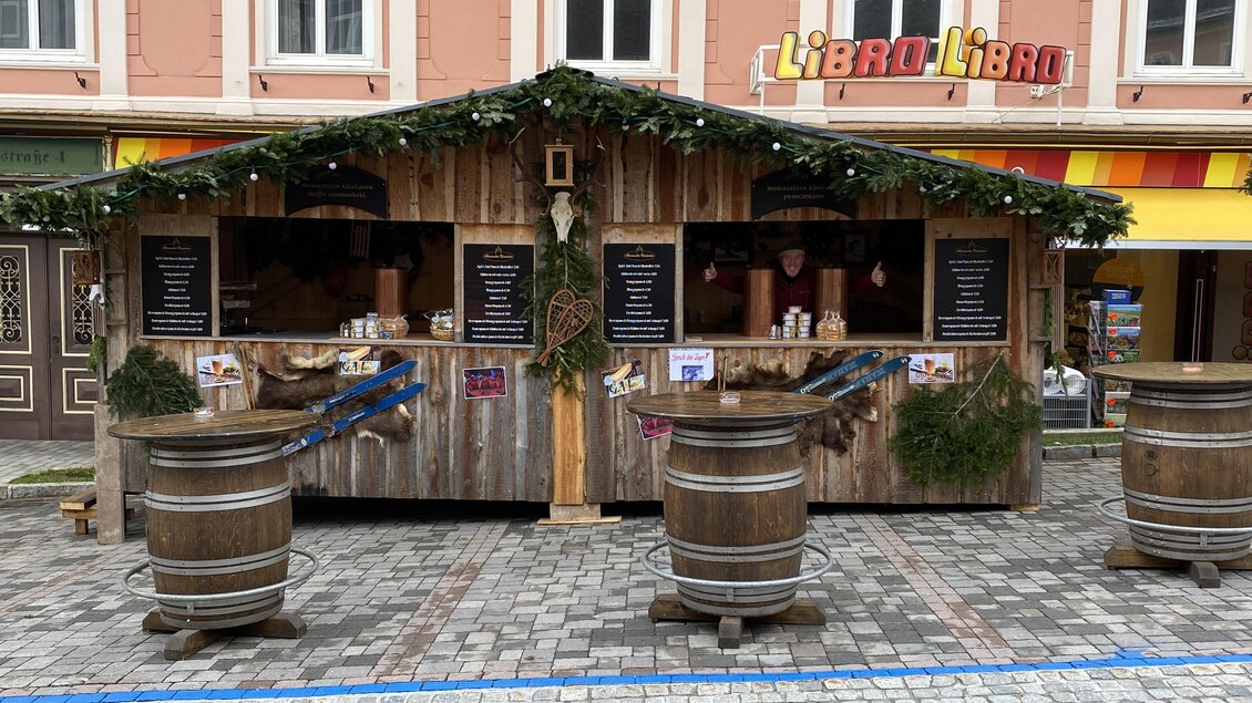 Ein schön dekorierter Weihnachtsstand aus Holz mit Fichtenzweigen. Im Hintergrund sind bunte Ladenfronten zu sehen.