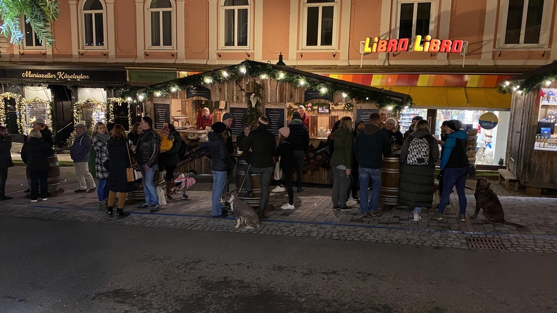 Ein belebter Weihnachtsmarkt mit Menschen, die an Ständen stehen. Die Umgebung ist festlich beleuchtet.