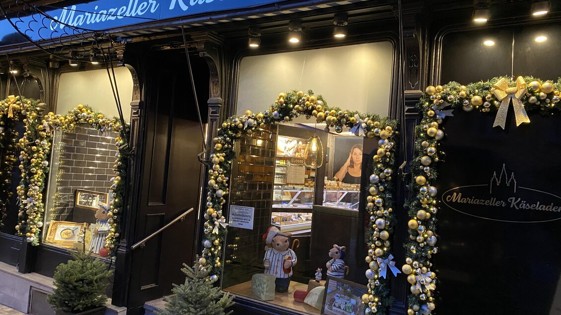 Ein Käseladen mit festlicher Dekoration und Lichtern. Die Fenster sind geschmückt und laden zum Einkaufen ein.