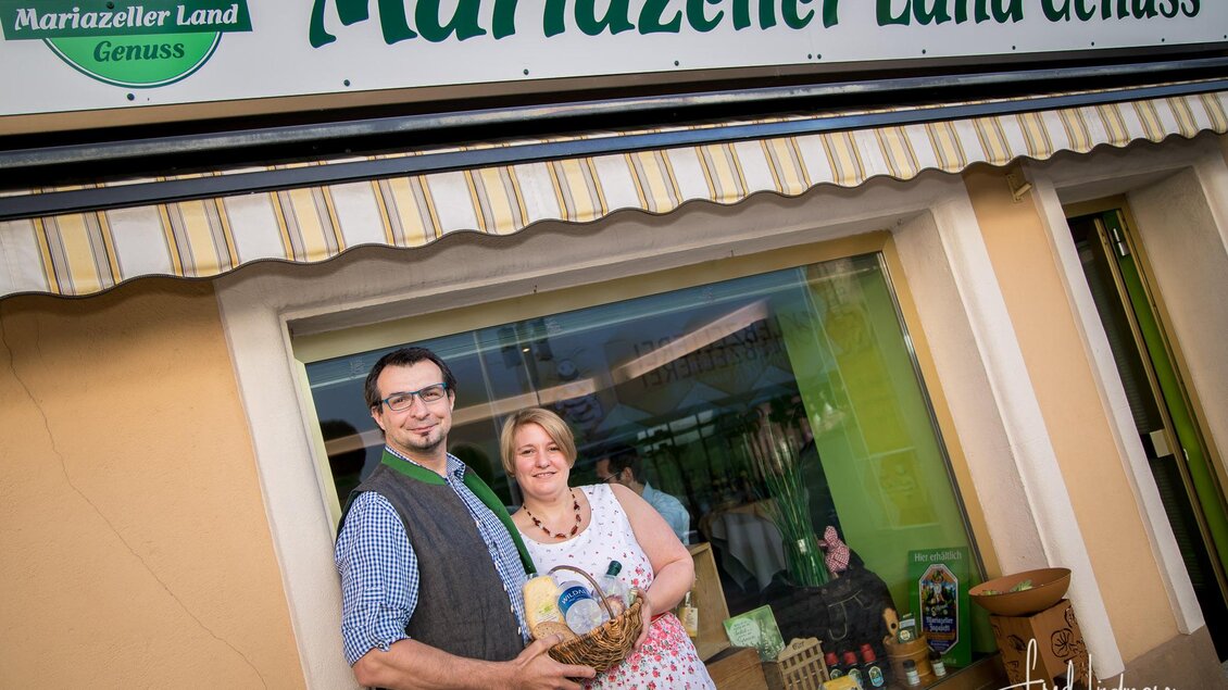 Ein Paar steht vor einem Geschäft namens "Mariazeller Land Genuss". Sie halten einen Korb und lächeln freundlich in die Kamera. | © Fred Lindmoser