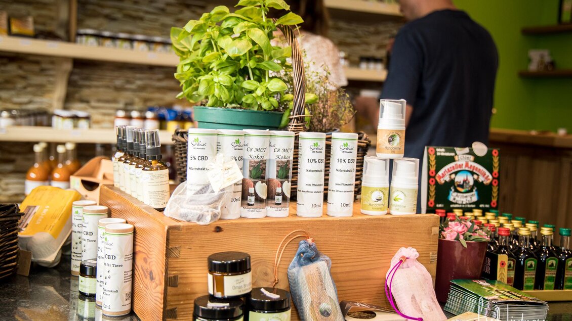 Ein Verkaufsstand mit verschiedenen Naturprodukten, darunter Kräuter, Öle und Flaschen. Im Hintergrund sind Menschen zu sehen, die die Produkte durchstöbern. | © Fred Lindmoser