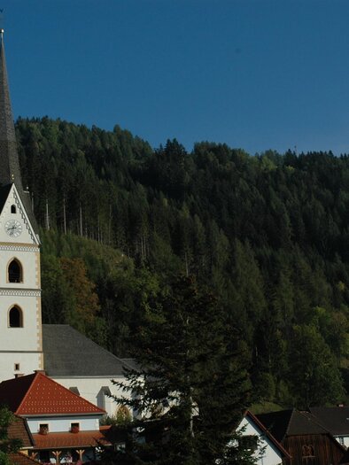 A picturesque church with a pointed tower stands in front of a hilly forest. The surroundings are green and idyllic. | © Tourismusverband Murau