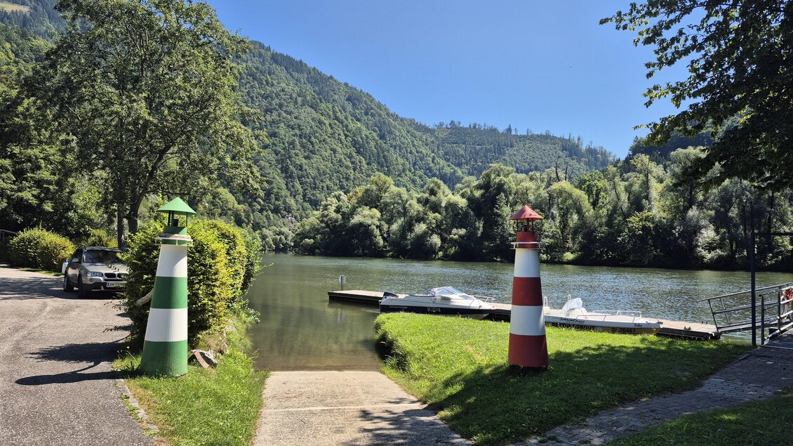 Ein ruhiger Fluss mit grünen Hügeln im Hintergrund. Auf beiden Seiten des Weges stehen farbige Leuchttürme. | © Andrea Stelzer