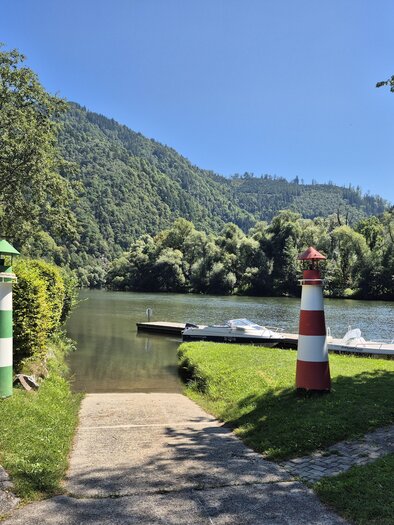 Ein ruhiger Fluss mit grünen Hügeln im Hintergrund. Auf beiden Seiten des Weges stehen farbige Leuchttürme. | © Andrea Stelzer
