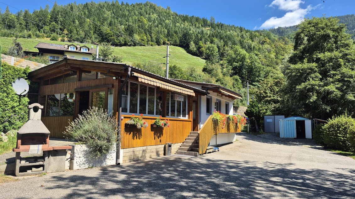 Ein gemütliches Holzhaus umgeben von Bäumen und Hügeln. Im Vordergrund ist ein Weg und ein Grillplatz zu sehen. | © Andrea Stelzer