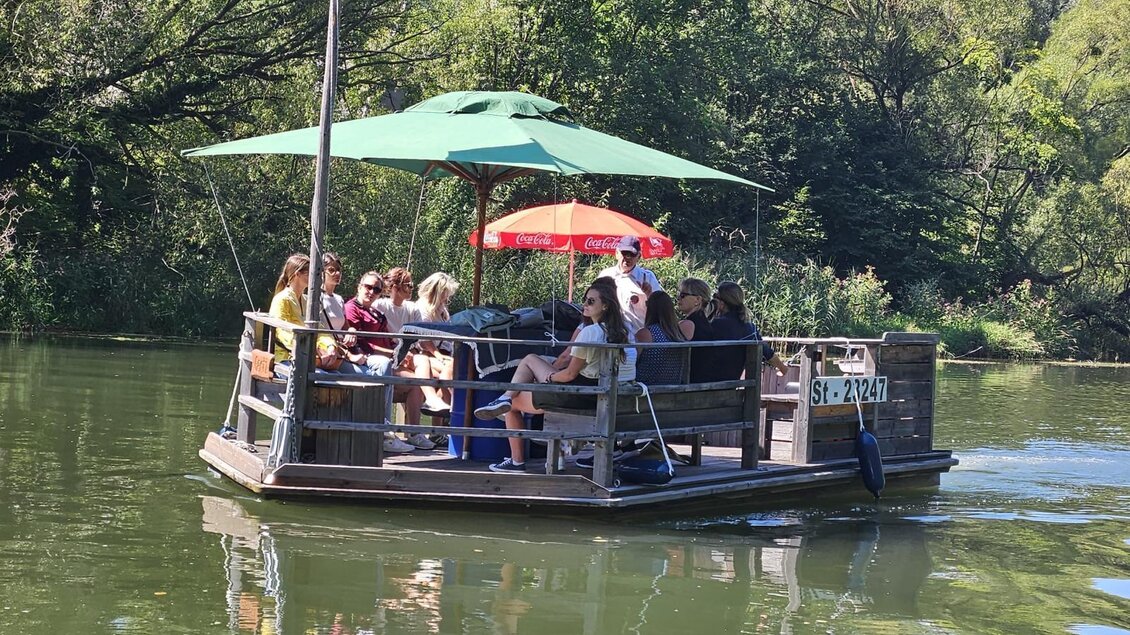 Eine Floßfahrt auf einem ruhigen Gewässer mit mehreren Menschen. Das Floß hat ein Sonnendach und ist von grünen Bäumen umgeben. | © Andrea Stelzer