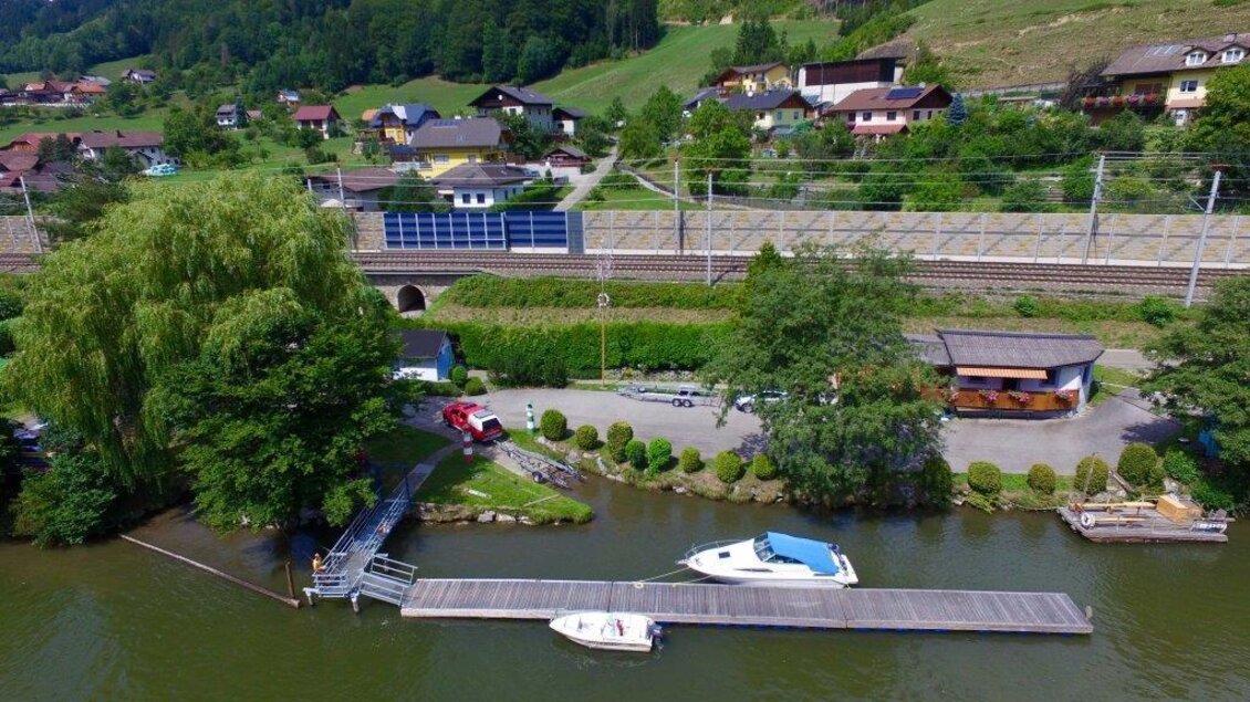 Ein malerischer See mit einem Steg und einem kleinen Boot. Im Hintergrund sind grüne Hügel und Wohnhäuser zu sehen. | © Prinz Eugen
