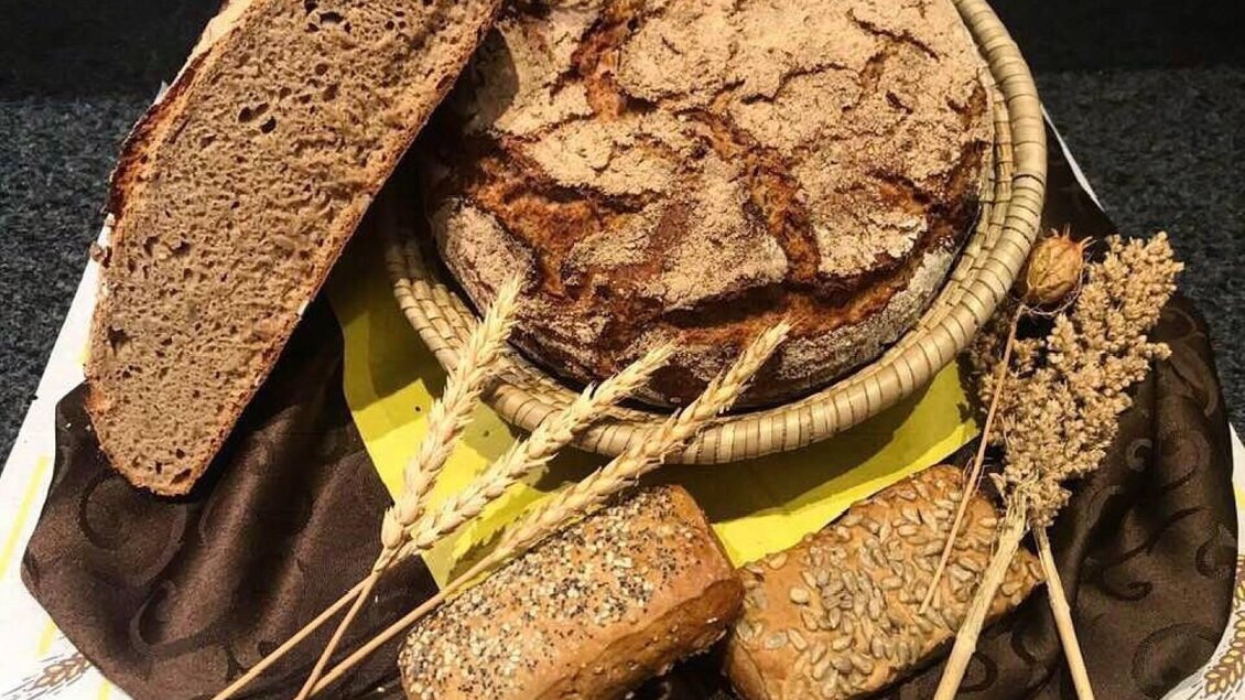 Ein rustikaler Brotkorb mit verschiedenen Brotsorten, darunter ein großes Bauernbrot und kleine Brötchen. Dekorative Getreideähren ergänzen das Arrangement. | © Marktcafé Wildbacher