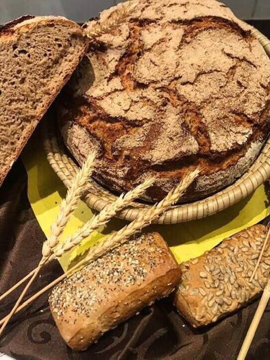 A rustic bread basket with various types of bread, including a large country loaf and small rolls. Decorative ears of grain complement the arrangement. | © Marktcafé Wildbacher