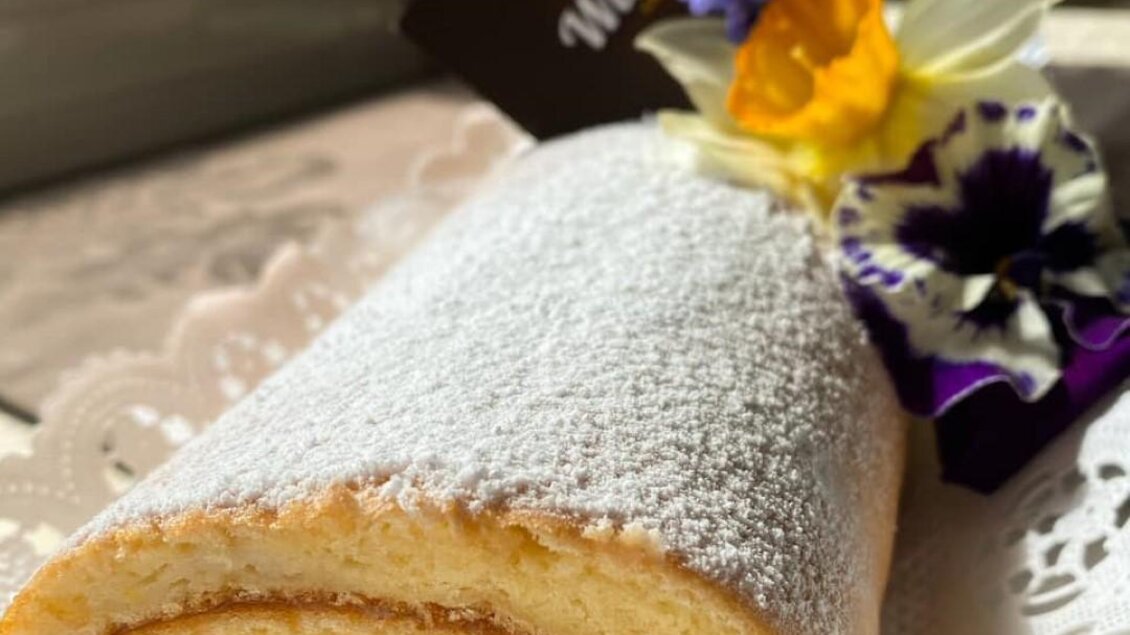 Eine frisch gebackene Biskuitrolle mit Puderzucker bestäubt. Dekoriert mit essbaren Blüten und einer eleganten Markenkarte. | © Marktcafé Wildbacher