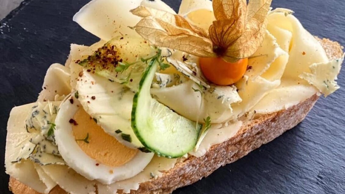 Ein belegtes Brot mit Käsescheiben, einer Scheibe Ei und frischem Gemüse. Oben ist eine essbare Blüte und eine kleine Tomate dekoriert. | © Marktcafe Wildbacher