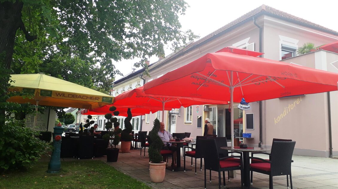 Ein Restaurant mit roten Sonnenschirmen draußen. Die Gäste sitzen an Tischen und genießen die Atmosphäre. | © Marktcafé Wildbacher
