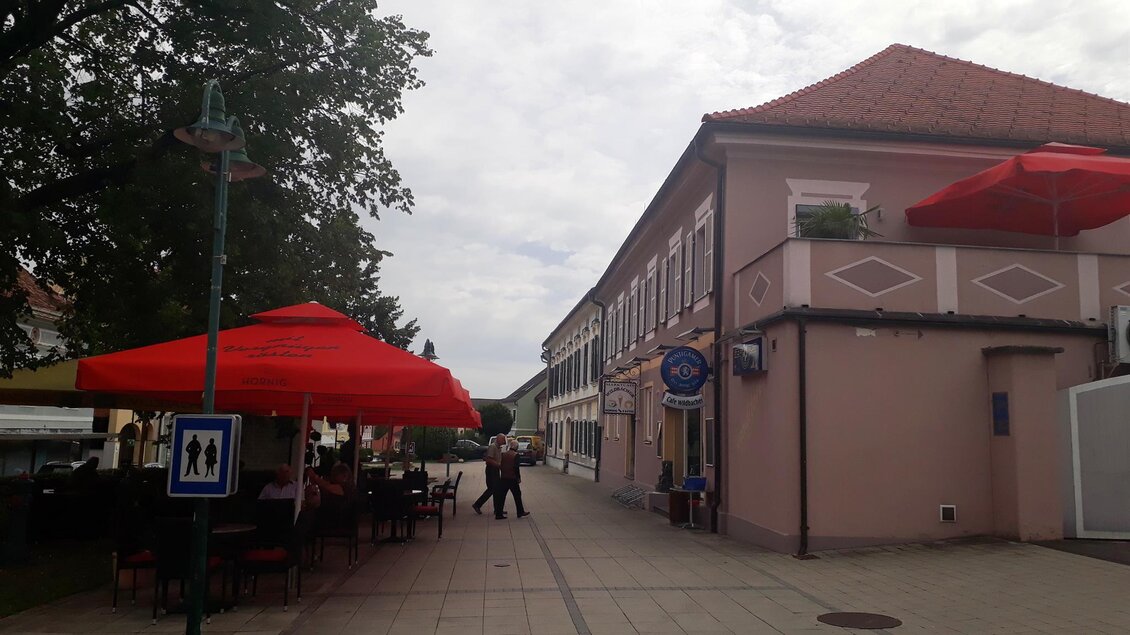 Eine ruhige Straße mit Cafés und roten Sonnenschirmen. Zu sehen sind eine Reihe von Gebäuden und eine angenehme Atmosphäre. | © Marktcafé Wildbacher