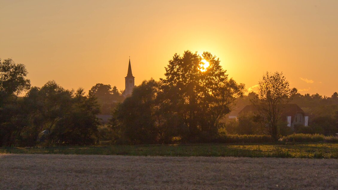 Ein wunderschöner Sonnenuntergang über einer ländlichen Landschaft. Im Hintergrund sind Bäume und ein Kirchturm zu sehen. | © Sandra Brünner