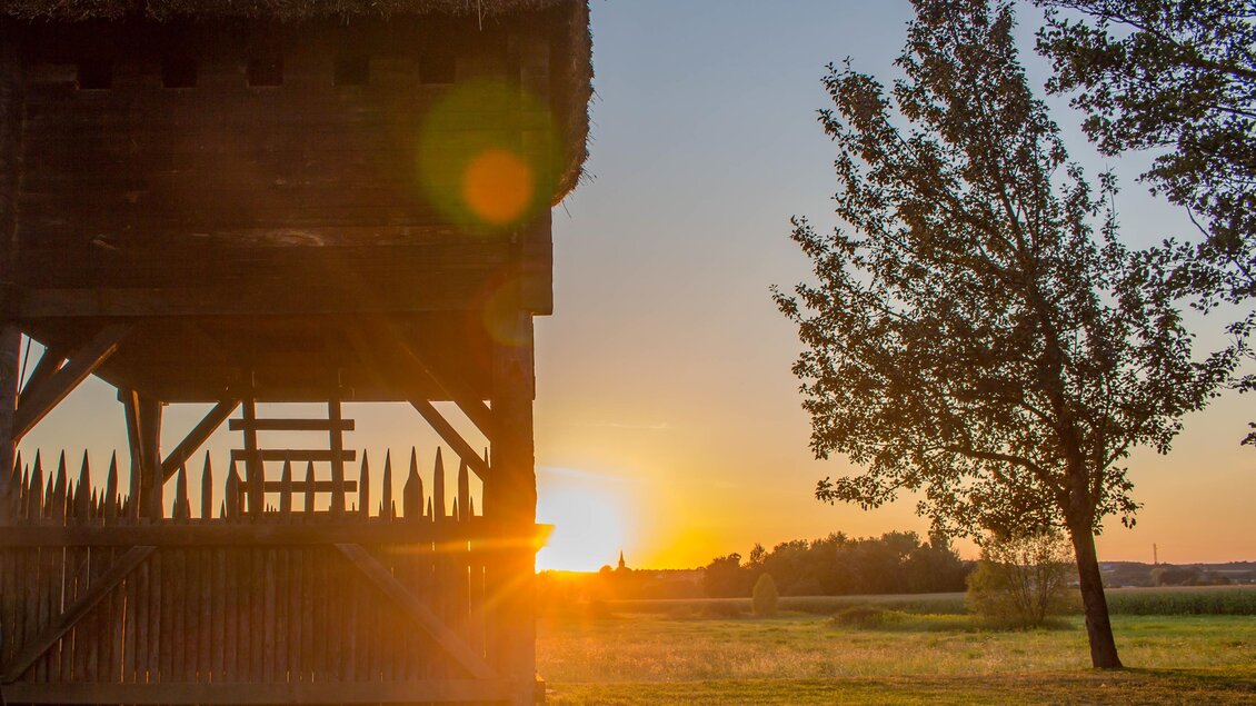 Ein traditionelles Holzgebäude mit Strohdach, umgeben von Bäumen. Im Hintergrund geht die Sonne unter und beleuchtet die Landschaft. | © Sandra Brünner