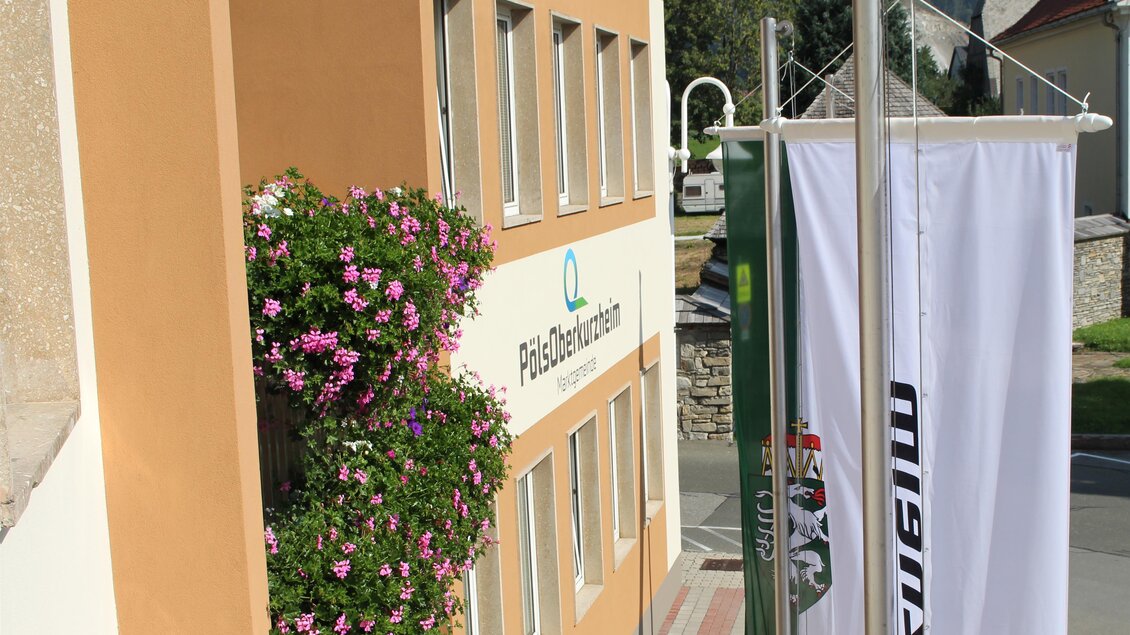 Ein helles Gebäude mit einem Blumenkasten voller pinker Blumen. Eine Fahne ist neben dem Eingang aufgestellt. | © Marktgemeinde Pöls-Oberkurzheim