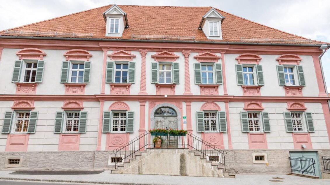 Ein historisches Gebäude mit rosa Anstrich und grünen Fensterläden. Es hat ein schickes Eingangsportal und einen klaren Himmel im Hintergrund. | © Marktgemeinde Wies