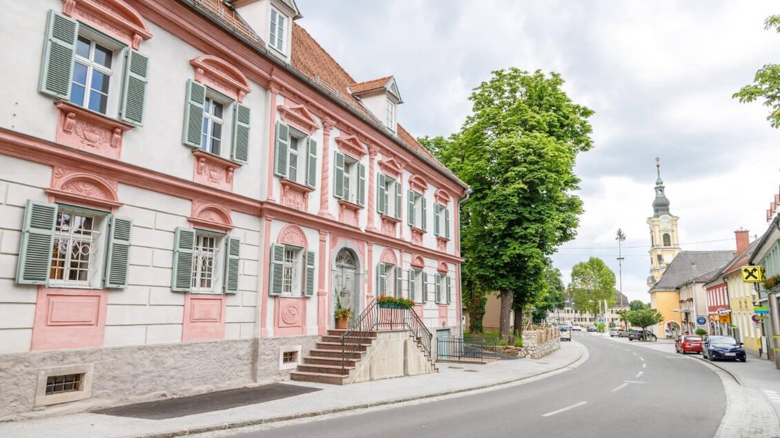 Ein historisches Gebäude mit rosa Fassaden und grünen Fensterläden. Im Hintergrund ist ein Kirchturm und eine ruhige Straße zu sehen. | © Marktgemeinde Wies