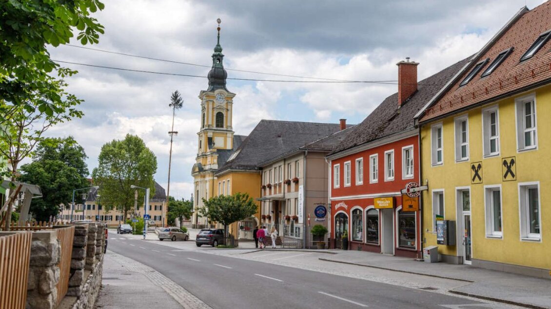 Eine ruhige Straße mit bunten Häusern und einem Kirchturm im Hintergrund. Der Himmel ist bewölkt und es sind einige Bäume entlang der Straße zu sehen. | © Marktgemeinde Wies