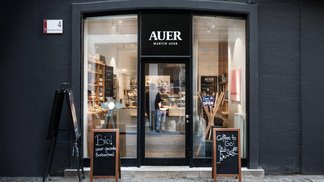 Einladendes Geschäft mit dem Namen "Auer" und großen Fenstern. Vor dem Eingang stehen Schiefertafeln mit Angeboten und Informationen. | © Martin Auer