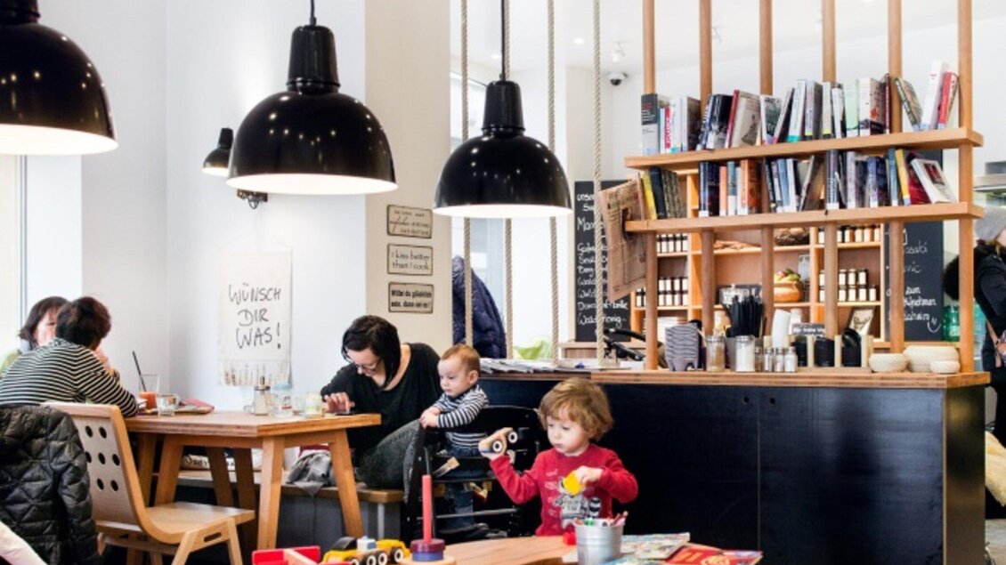 Ein modernes Café mit Holztischen und Schwarz-Weiß-Lampe. Eine Familie genießt die Zeit zusammen, während ein Kind mit Spielzeug spielt. | © Martin Auer