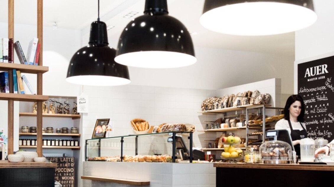 Ein modernes Café mit einer einladenden Theke und frischen Backwaren. Große Lampen hängen von der Decke und eine Barista bereitet Getränke zu. | © Martin Auer