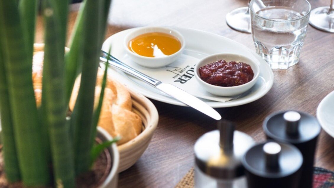 Ein Tisch mit einer Auswahl an Dips, darunter Honig und eine Sauce, sowie frischen Brötchen. Im Hintergrund sind Pflanzen und Wassergläser zu sehen. | © Martin Auer