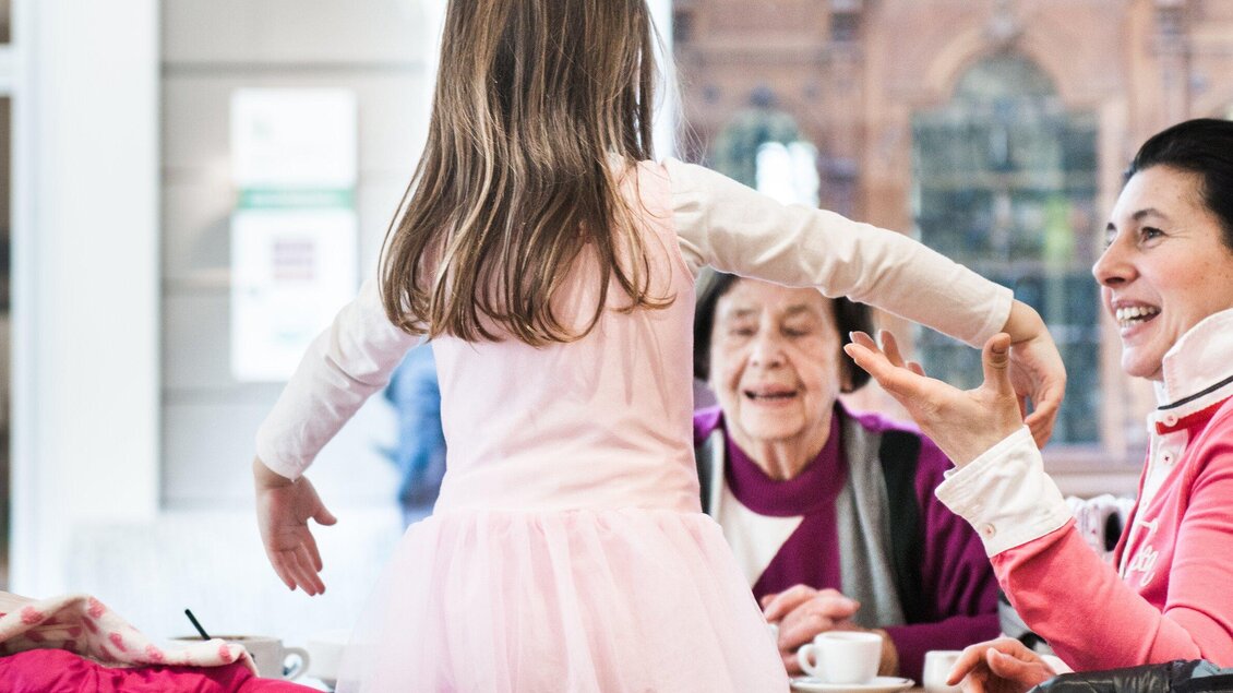 Ein Mädchen in einem rosa Kleid tanzt fröhlich, während zwei Erwachsene am Tisch sitzen und zuschauen. Die Atmosphäre ist freundlich und einladend. | © Martin Auer