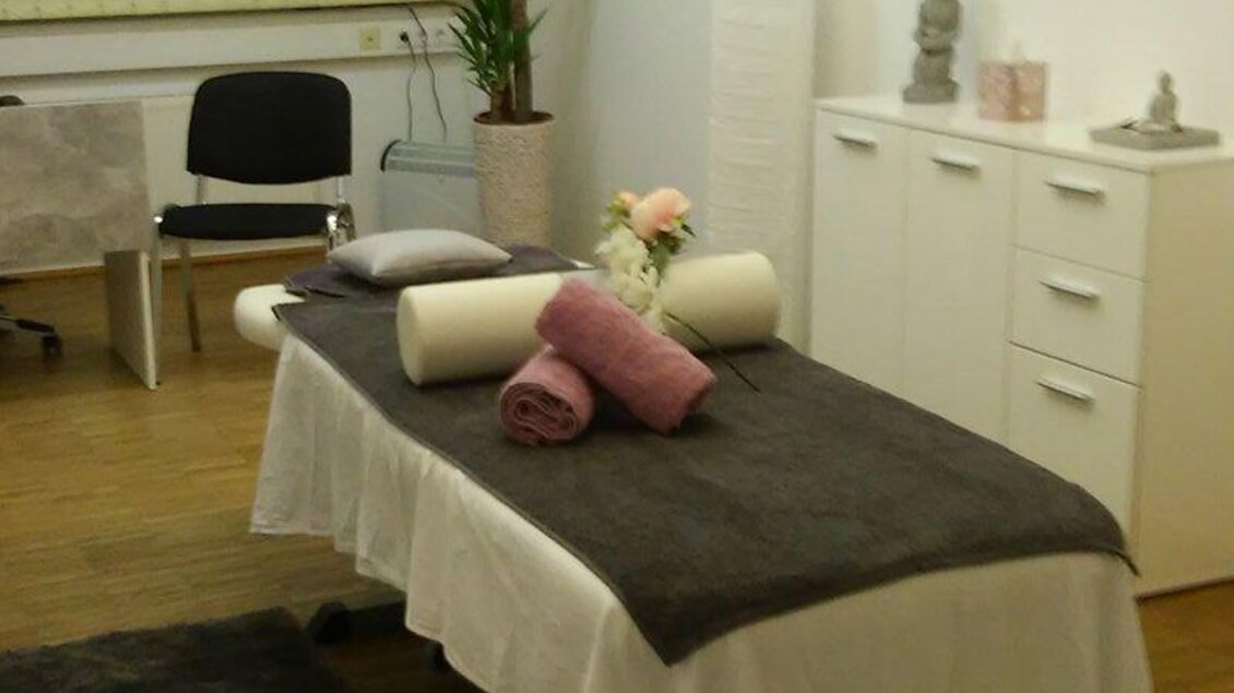 Ein ruhiger Massagebereich mit einem Behandlungstisch, dekoriert mit Handtüchern und Blumen. Pflanzen und ein grauer Teppich sorgen für eine entspannte Atmosphäre.