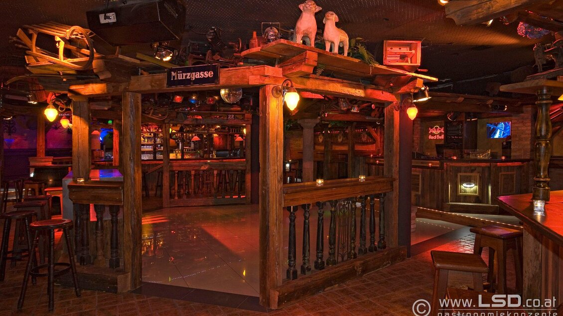 Eine gemütliche Bar mit Holzdetails und sanfter Beleuchtung. Der Raum hat eine rustikale Atmosphäre mit Blick auf die Theke und Sitzbereiche. | © Mausefalle