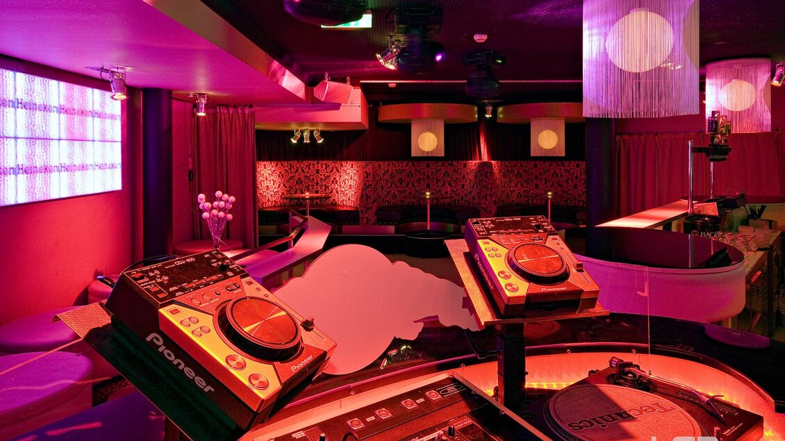 Ein stilvoller Nachtclub mit rot beleuchtetem Ambiente und DJ-Pulten. Weiche Sofas und moderne Dekorationen schaffen eine einladende Atmosphäre. | © Mausefalle
