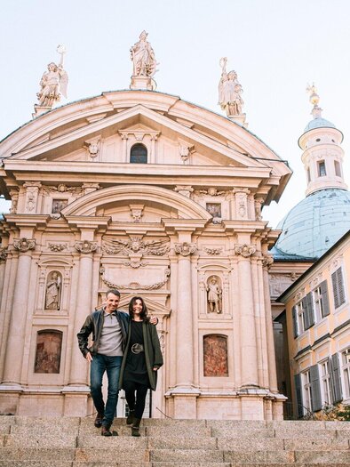 Ein Paar steht auf einer Treppe vor einem historischen Gebäude mit kunstvollem Fassadendekor. Die Umgebung zeigt enge Gassen und bunte Gebäude im Hintergrund. | © Graz Tourismus