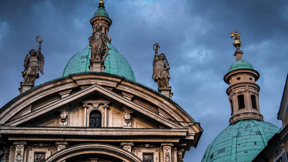 Eine eindrucksvolle Kirchenarchitektur mit einer grünen Kuppel und grazilen Statuen. Der Himmel ist bewölkt und verleiht der Szene eine dramatische Stimmung.