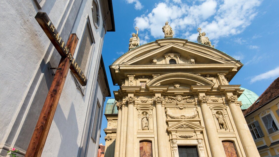 Eine beeindruckende Kirche mit aufwendiger Fassade und Skulpturen. Neben der Kirche steht ein großes Holzkreuz. | © Graz Tourismus - Harry Schiffer
