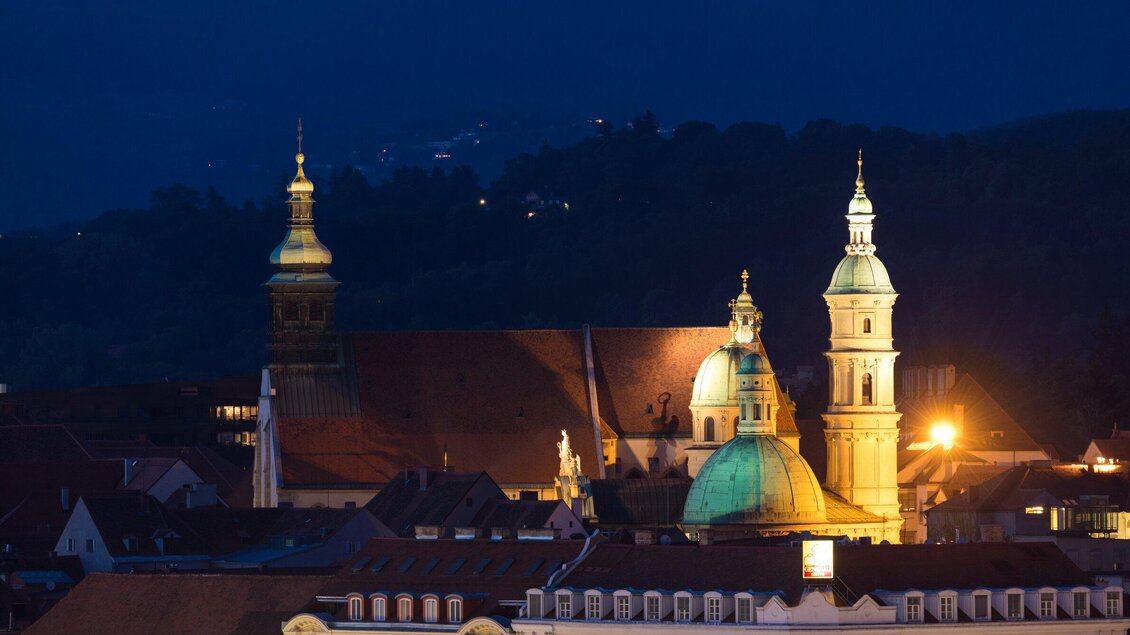Eine nächtliche Stadtansicht mit beleuchteten Kirchenkuppeln. Der Himmel ist dunkelblau, während die Lichter der Stadt warme Akzente setzen. | © Graz Tourismus - Harry Schiffer
