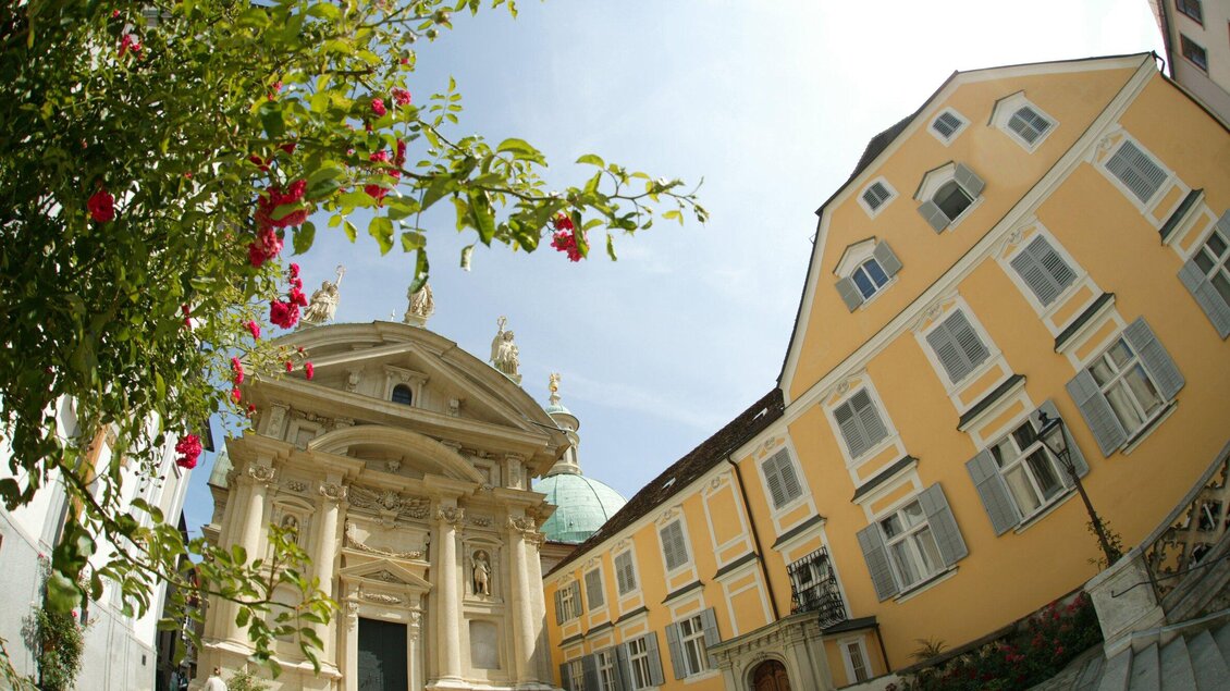 Eine historische Kirche mit einer kunstvollen Fassade und einer Statue auf dem Dach. Im Vordergrund wachsen rosa Blumen, und im Hintergrund sieht man bunte Gebäude. | © Graz Tourismus - Hans Wiesenhofer